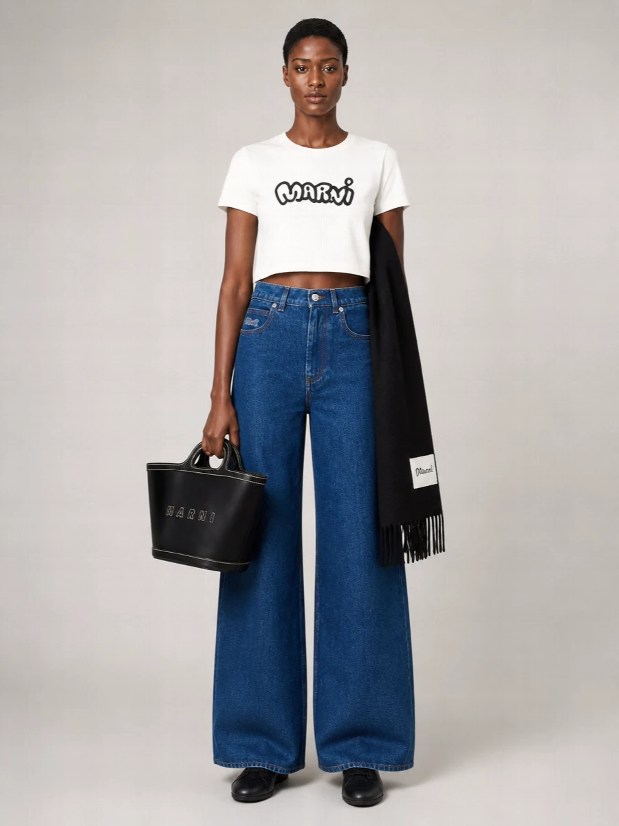 A cropped logo tee tops rigid wide-leg indigo denim for a clean, graphic take on streetwear that still reads grown-up. Sleek black sneakers and a structured black bucket bag keep the lines crisp, while a long black scarf adds depth and subtle branding without diluting the monochrome punch.