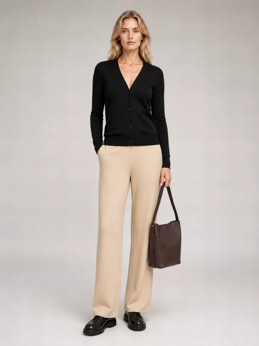 A black V-neck cardigan sits against fluid light camel trousers for a soft, vertical line in quietly neutral tones. Chunky black derbies add structure without heaviness, while a chocolate leather bucket bag deepens the palette with restrained richness. The result is relaxed but composed, ideal for understated days in the city or a low-key dinner out.