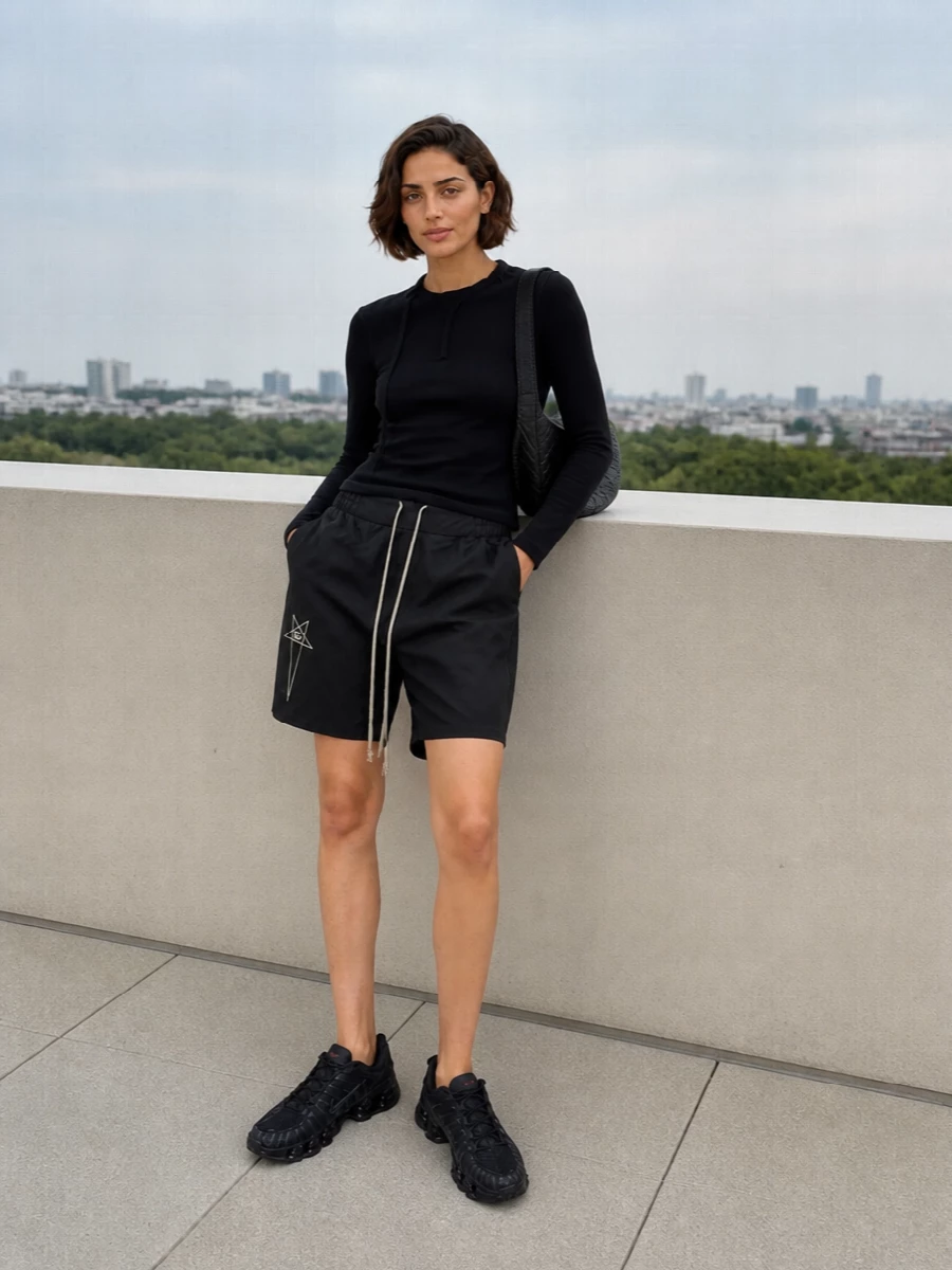 A sharp, monochrome base lets the Rick-inspired shorts lead, paired with a close-cut black ribbed top that keeps everything lean above the knee. Technical black runners echo the sport DNA without breaking the palette, while the sculpted shoulder bag adds a subtle architectural contrast to the relaxed volume of the shorts. The result is a clean, directional street outfit that feels ready for city movement rather than gym time.