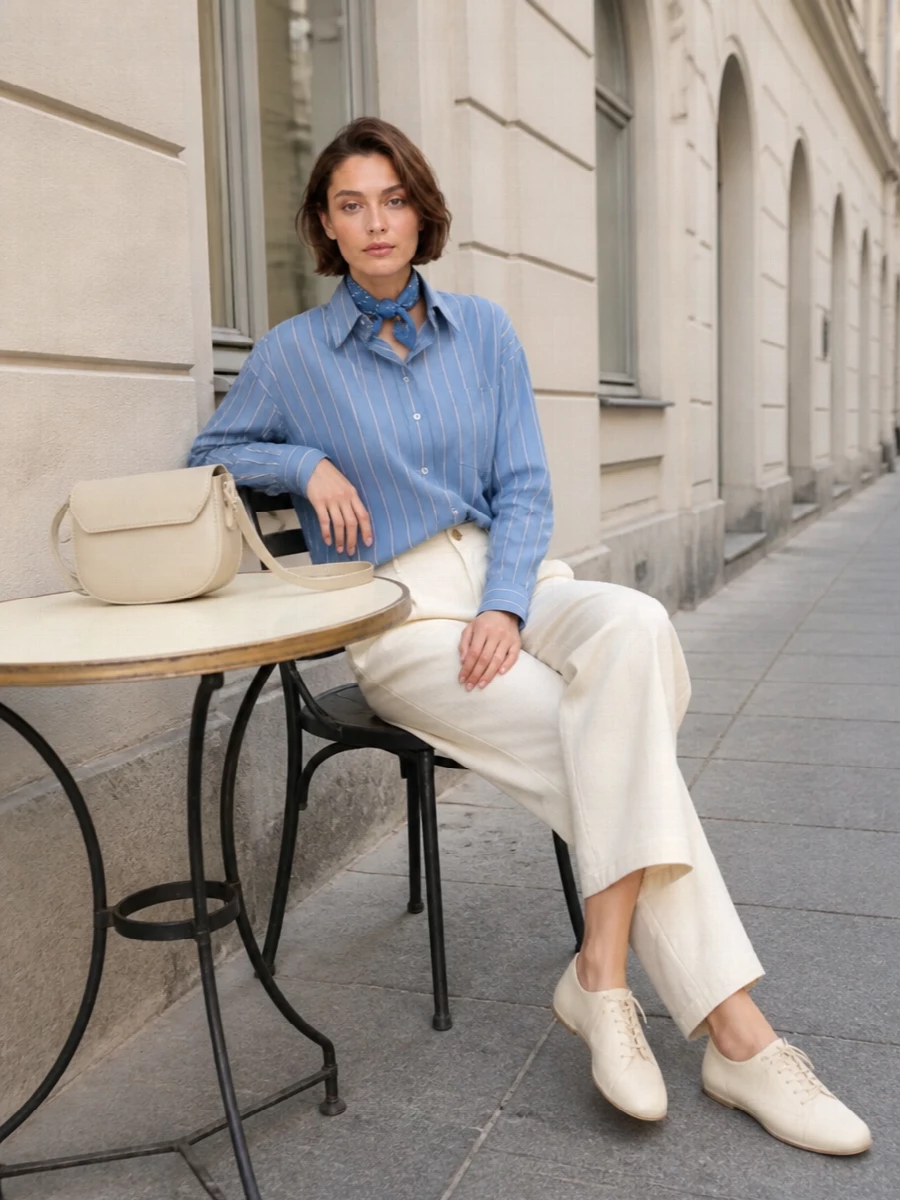 An oversized striped shirt meets soft pleated trousers for a relaxed, borrowed-from-him silhouette that still feels sharp. Buttercream leather accessories keep the palette quiet and coherent, while a chalky blue scarf adds a small, graphic twist. The result is polished enough for the office yet easy for a long city day.