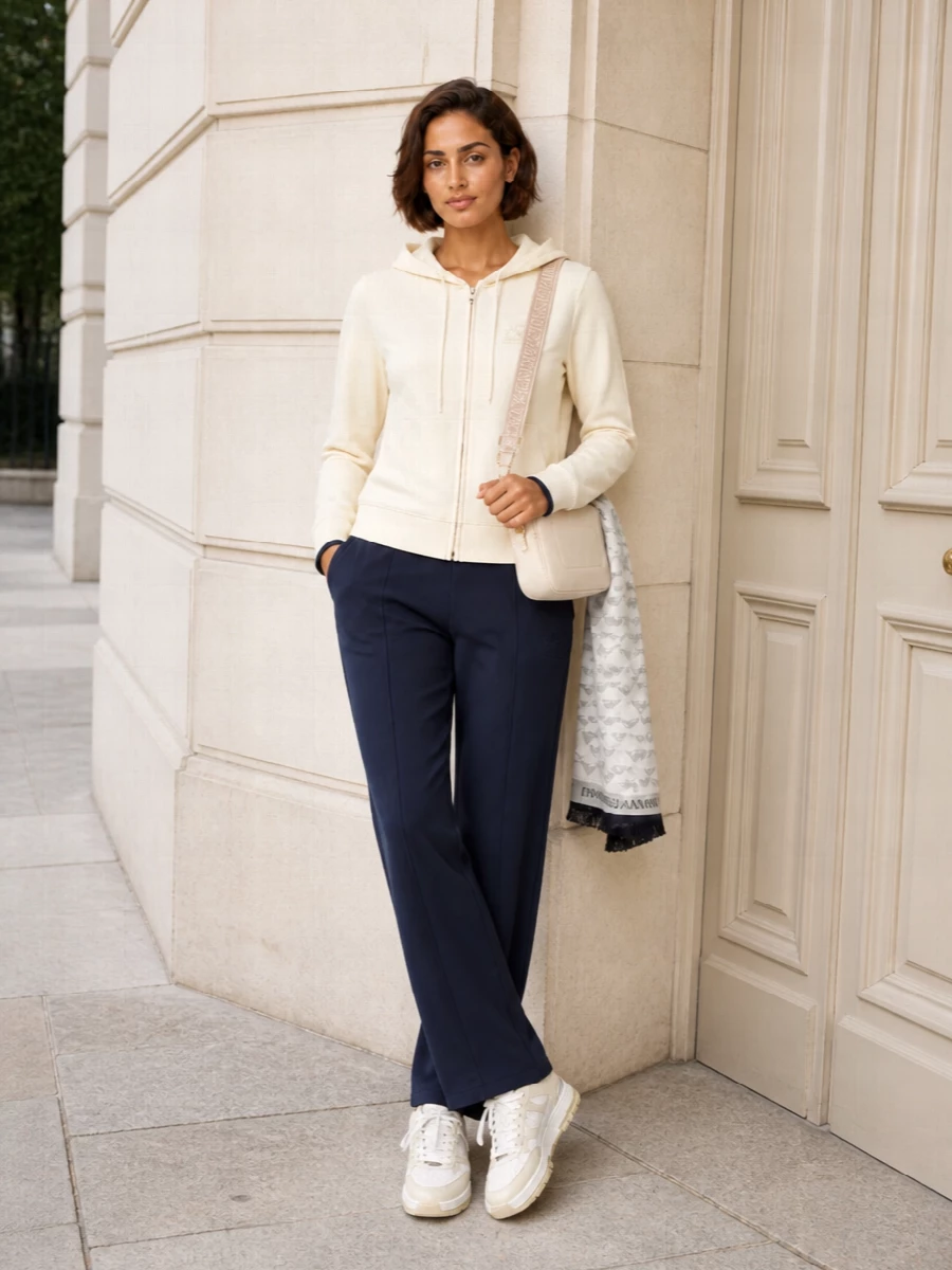 Warm ivory on top and in the accessories is grounded with clean navy straight-leg joggers, keeping the set quietly sporty and minimal. Off-white court sneakers keep everything relaxed yet considered, while the compact cross-body adds just enough structure for city errands and travel days. The tonal scarf introduces a soft layer of texture without breaking the calm, neutral palette.