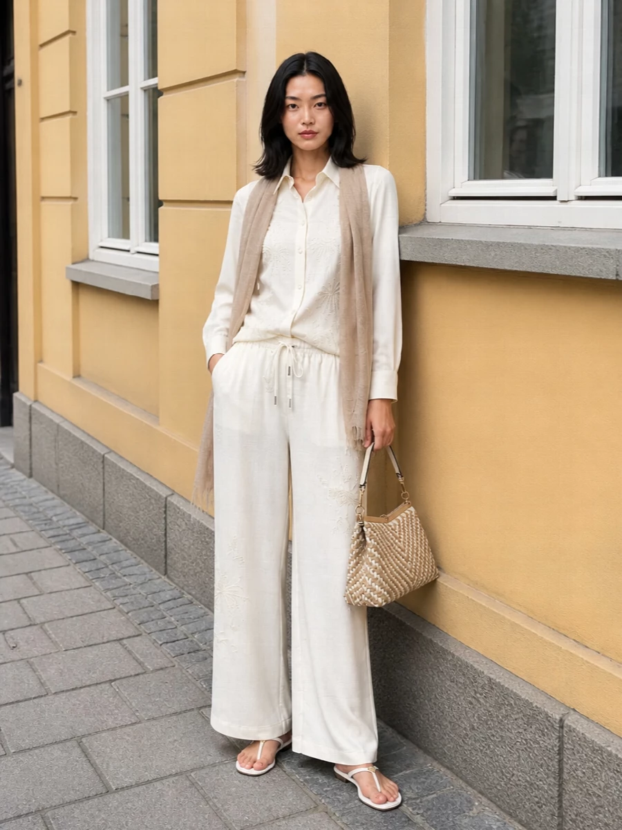 An all‑neutral story that lets Ermanno Scervino’s embroidery do the talking, this pairing keeps everything light, fluid and quietly polished. The regular ivory shirt tucks into the wide‑leg trouser to create a soft column, while barely‑there white sandals and a woven top‑handle bag add just enough texture for terrace lunches or coastal afternoons. A whisper‑light sand scarf finishes the outfit with a barely-there hint of warmth and movement.