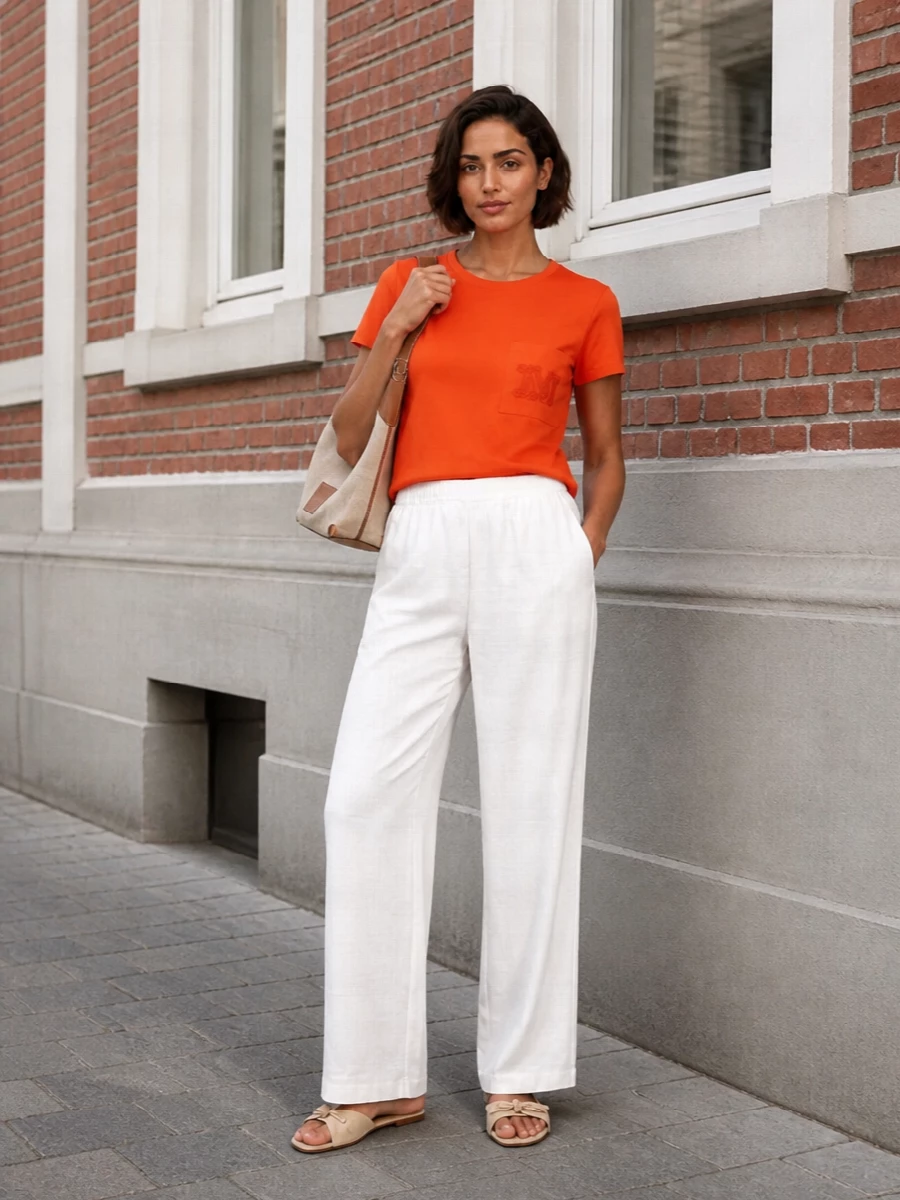 A vivid orange tee is grounded with soft white wide-leg trousers for a clean, modern take on relaxed day dressing. Butter-toned slides and a canvas shoulder bag keep the palette warm and neutral, letting the color pop stay focused on the top. The result is quietly polished and easy for city days, coffee runs, or travel when you still want the outfit to feel intentional.