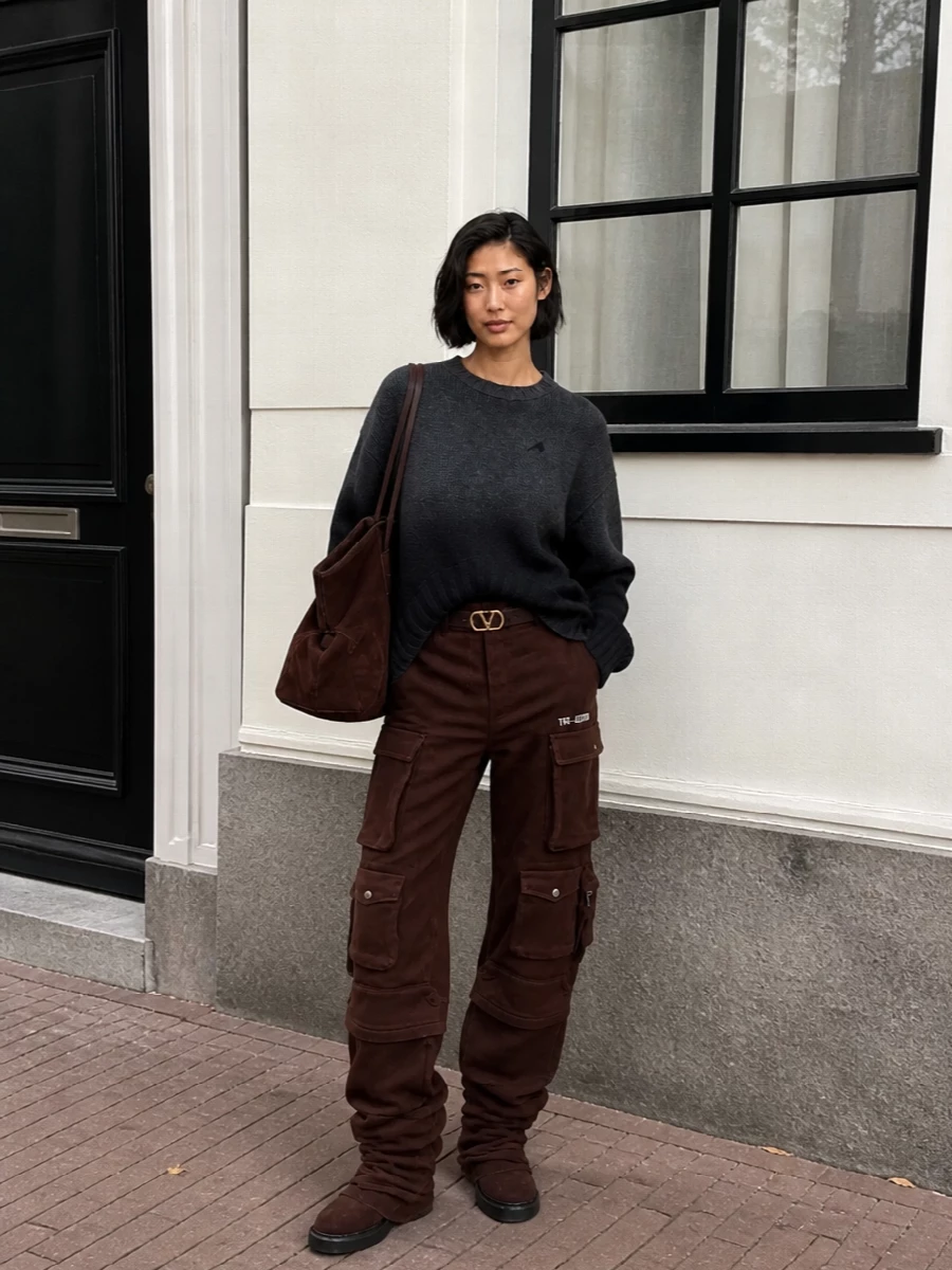 Deep brown cargos, a charcoal ombré knit and matching slouchy suede boots create a low-key, tonal city uniform with real attitude. The oversized suede tote keeps the proportions intentionally generous, while a slim chocolate logo belt hints at structure without breaking the relaxed line. It is plush, grounded streetwear that still reads unmistakably designer.
