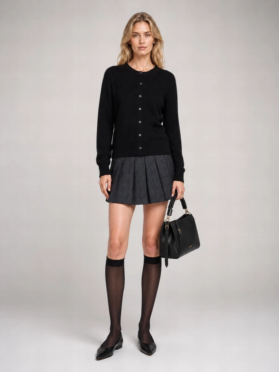 Dunst’s black cardigan and checked pleated mini skirt create a studied, campus-leaning uniform in shades of charcoal and black. Sheer knee-highs and pointed flats keep the line long and neat, while the structured black shoulder bag adds just enough everyday polish. The result is quietly preppy, made for libraries, galleries and unhurried city days.