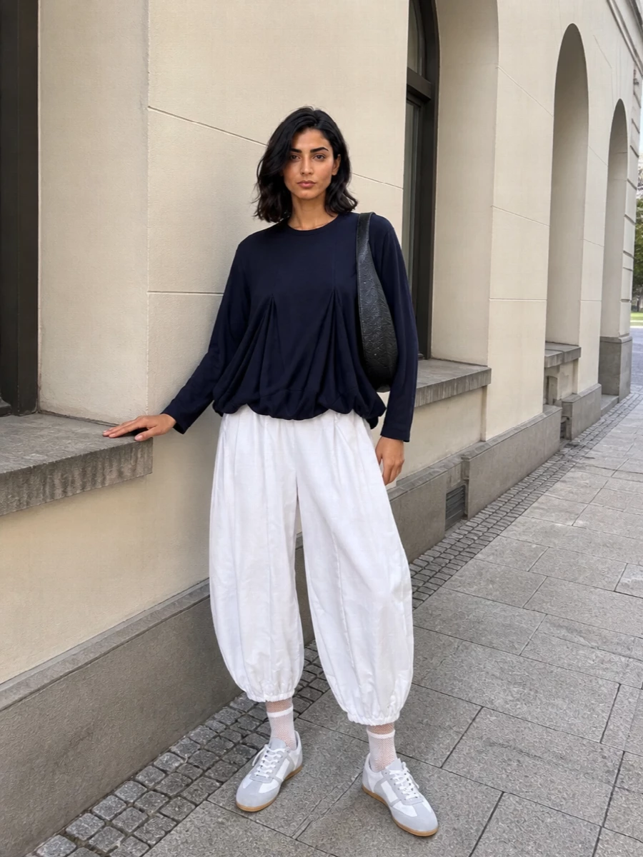 Architectural volume on top and bottom creates a cocooned silhouette, with navy balancing the bright white trouser for a clean, graphic feel. Soft grey-white sneakers and a sculpted black shoulder bag keep the palette tight and the mood urban, so the proportions stay experimental but wearable for daytime. Mesh socks add just enough texture to make the details feel considered rather than cute.