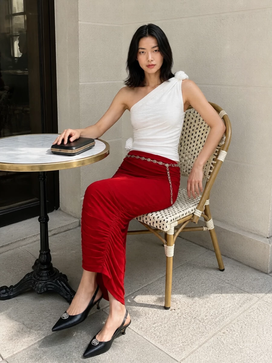 A deep red ruched column skirt meets a sculpted white one-shoulder top for a romantic, modern cocktail pairing that feels sensual yet precise. Black kitten-heel slingbacks and a hard-case clutch ground the color story, keeping the focus on drape and silhouette. A delicate silver chain belt traces the waist, adding a whisper of shine that moves with every step.