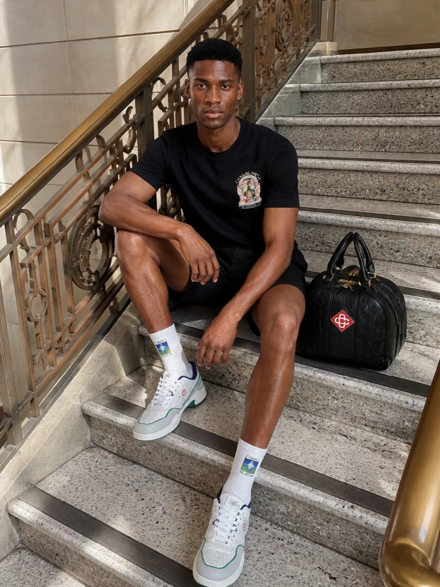 A black graphic tee sits over tailored jacquard shorts for a clean, city resort mood that still feels relaxed. White court-inspired sneakers and a structured embossed duffle keep the palette sharp and focused while leaning into Casablanca’s sport heritage. Ribbed striped socks add a small, playful color note that keeps the outfit fresh rather than flat monochrome.