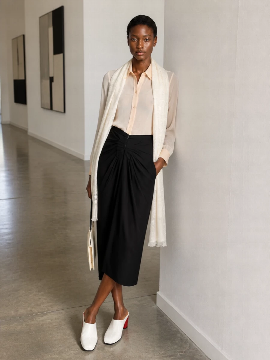 Sheer ivory and nude meet sculpted black in a quietly assured evening ensemble. The diaphanous shirt softens against the high-waisted draped midi, creating a long, refined line. Graphic white mules and a slim buttercream bag keep the palette calm while adding just enough edge for gallery nights or intimate dinners.