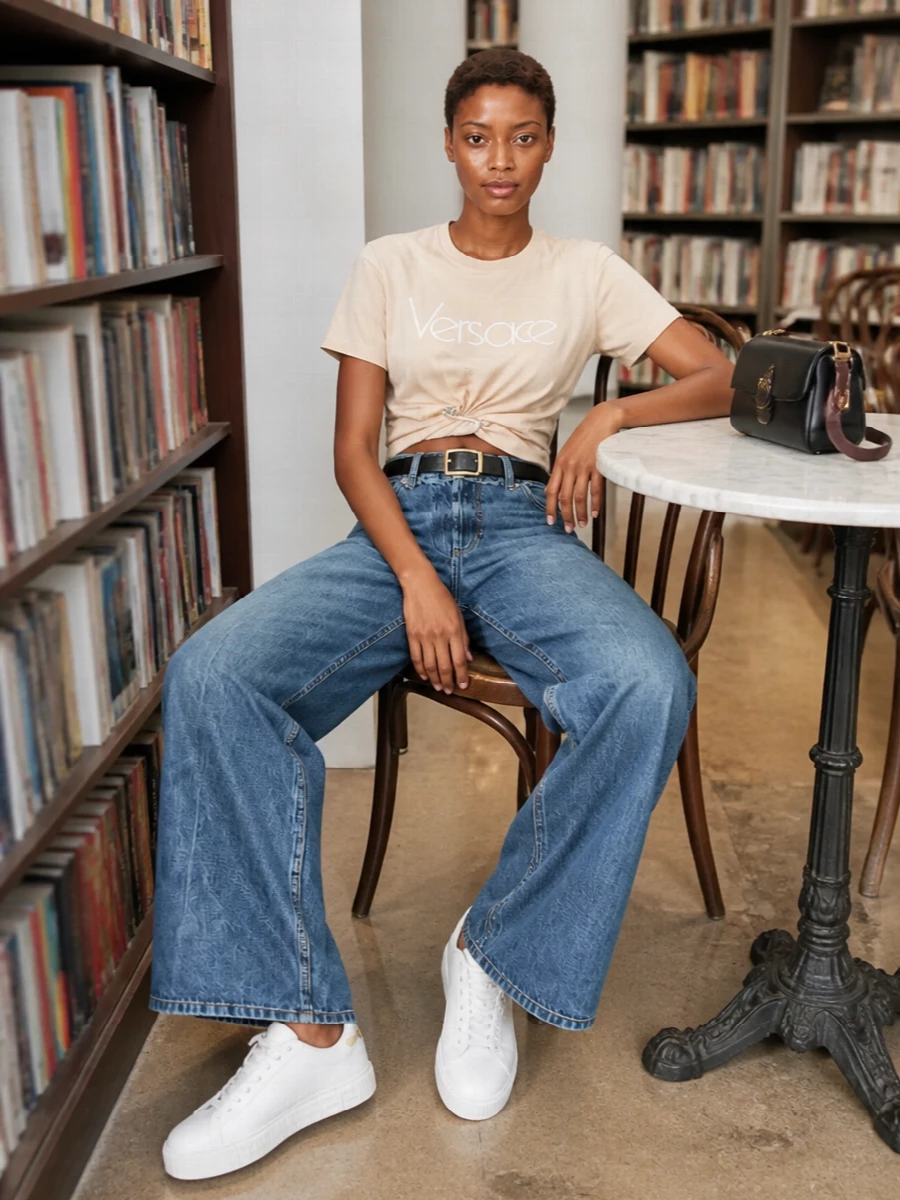 A sculpted beige logo tee meets clean mid-wash wide-leg denim for a minimal, street-led take on Italian polish. White leather sneakers keep the mood low-key, while a slim black shoulder bag and precise belt sharpen the finish just enough for city days and casual dinners. The result is easy, modern and quietly branded rather than logo-heavy.