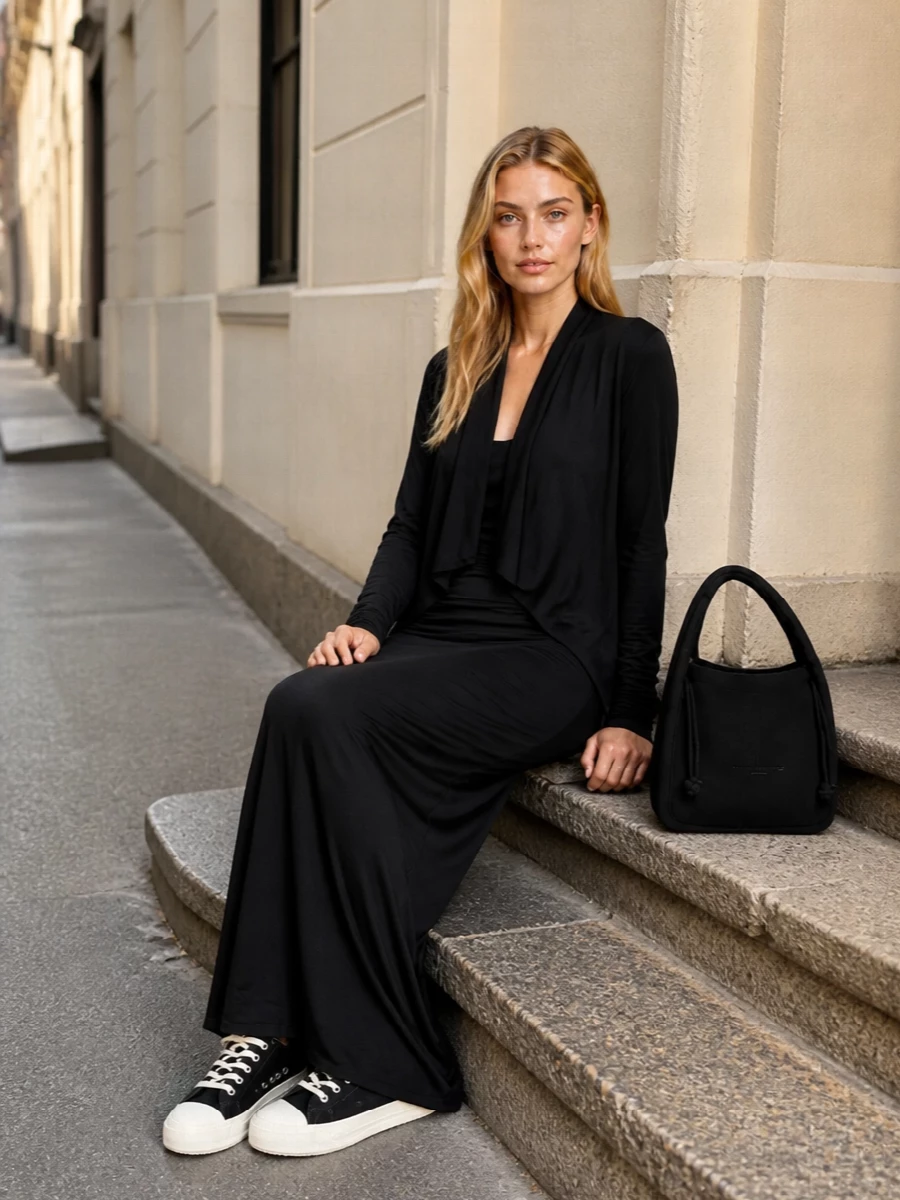 A long black knit column slips into a ruched jersey maxi, so the body reads as one continuous, fluid line. Graphic black and off-white sneakers cut the severity just enough, keeping the mood modern and grounded. A soft, curved shoulder bag in black completes the composition without interrupting the dark, minimalist flow.