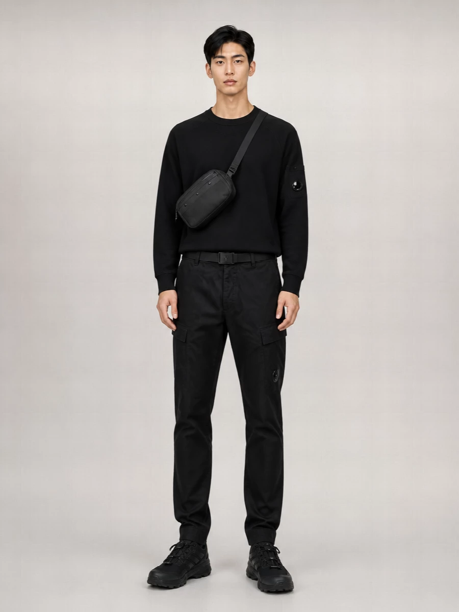 Head-to-toe black and charcoal keeps this C.P. Company cargo pairing sharp, letting the lens details and technical textures do the talking. The black crewneck and trail sneakers push it firmly into urban utility, practical for cold days without feeling heavy. A compact crossbody and low-profile belt finish the outfit with discreet function rather than extra noise.
