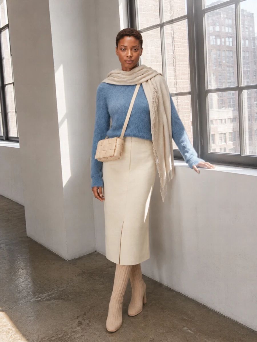 A dusty blue fuzzy crewneck meets a buttercream tailored midi skirt for a calm, precise take on winter texture. Ribbed knit knee boots and a compact woven cross-body hold the line clean and long, while an oatmeal wrap stays in the same hushed palette so everything feels quietly intentional. The result is soft to wear yet sharp enough for a polished day in the city.