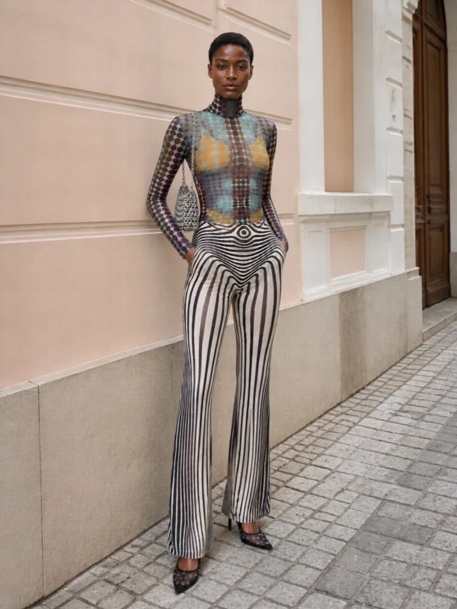 Gaultier’s trance-print mesh top meets his optic-stripe flares for a fully committed, second-skin evening statement that feels graphic rather than chaotic. Black mesh stilettos and a silver chainmail baguette sharpen the palette, keeping all the focus on the body-mapped prints and long, elongated lines. It is a club-first outfit that reads considered and directional from every angle.