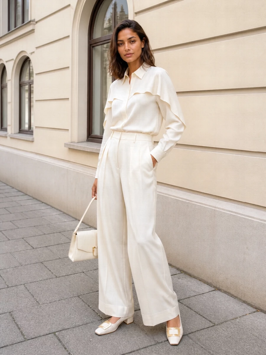 An ivory-on-ivory column keeps the focus on the blouse’s sculpted sleeve and clean placket, reading polished but softly romantic. High-waisted wide-leg trousers lengthen the line, while low block-heel slingbacks and a structured top-handle bag introduce just enough structure and shine for client meetings or dinner reservations. The blush-toned scarf is an optional touch of warmth that stays within the same quiet, neutral story.