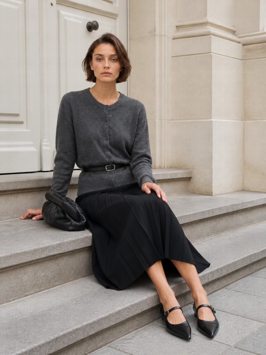 A black pleated midi, cinched with a slim belt and topped with a brushed charcoal cardigan, leans into a precise, collegiate uniform in a strictly monochrome palette. Pointed Mary Janes keep the mood grounded yet refined, while the sculpted pleated clutch quietly mirrors the skirt’s movement. The result is a polished, city-ready take on prep that is minimal, grown-up and quietly assured for cooler seasons.