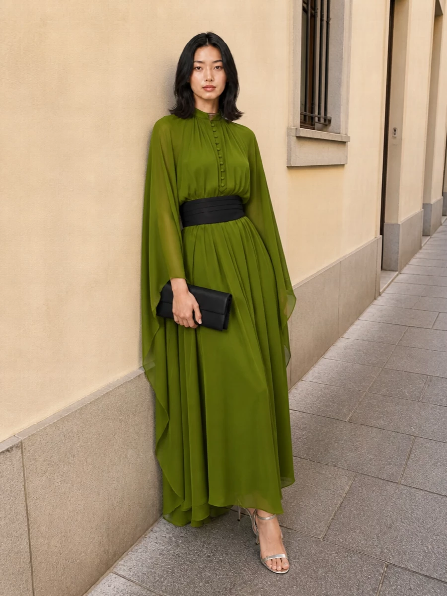 An olive Tom Ford caftan becomes sharply cinematic when anchored with a sculpted black cummerbund that defines the waist inside all that liquid volume. Silver ankle-strap stilettos and a slim black clutch keep the story precise and nocturnal, adding just enough metal to catch the light without competing with the dress.