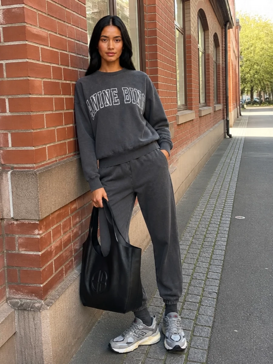 A washed-charcoal logo sweatshirt and matching-toned joggers create a monochrome, off-duty base that feels relaxed but intentional. Chunky grey runners keep it sporty and practical, while the sculpted black tote adds just enough structure for a city day. The result is a streamlined, model-off-duty uniform built around ease, texture and a tight grayscale palette.