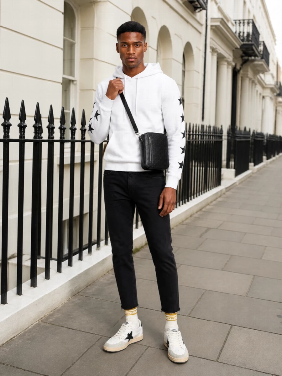 A clean black and white base keeps this Golden Goose hoodie in focus, with cropped faded-black denim sharpening the laid-back sport mood. Star-detailed sneakers and striped socks echo the hoodie’s graphics without feeling matchy, while a compact black crossbody adds just enough polish for city days or airport runs. The result is relaxed and athletic but still considered from head to toe.