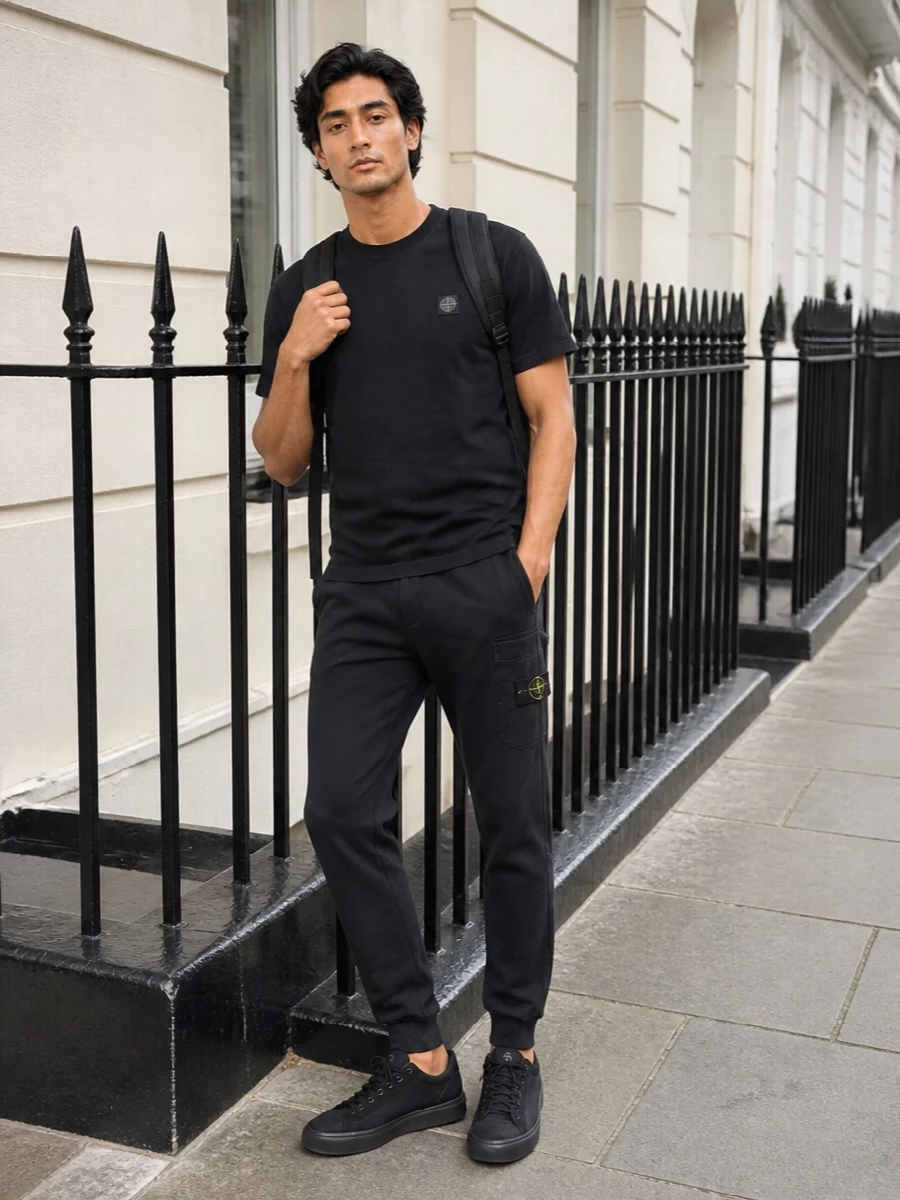 A washed-black Stone Island tee and matching tapered track pants create a tight, monochrome base that feels clean, athletic and quietly branded. All-black technical sneakers keep the line uninterrupted, while the structured backpack underlines the city-ready, movement-focused mood. The result is a minimal, logo-aware streetwear set that reads deliberate rather than loungey.