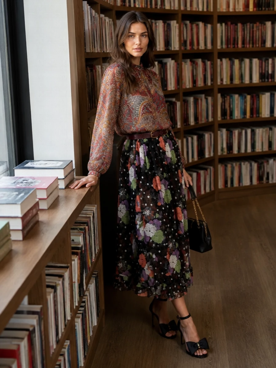 A sheer paisley blouse blurs into the floaty floral midi, creating a romantic mix of transparency and warm color over the graphic black base. A slim burgundy belt sharpens the high waist, while black and gold accessories keep the mood poised for cocktails rather than costume. The result is soft, bohemian movement anchored by quietly structured, city-ready finishing touches.