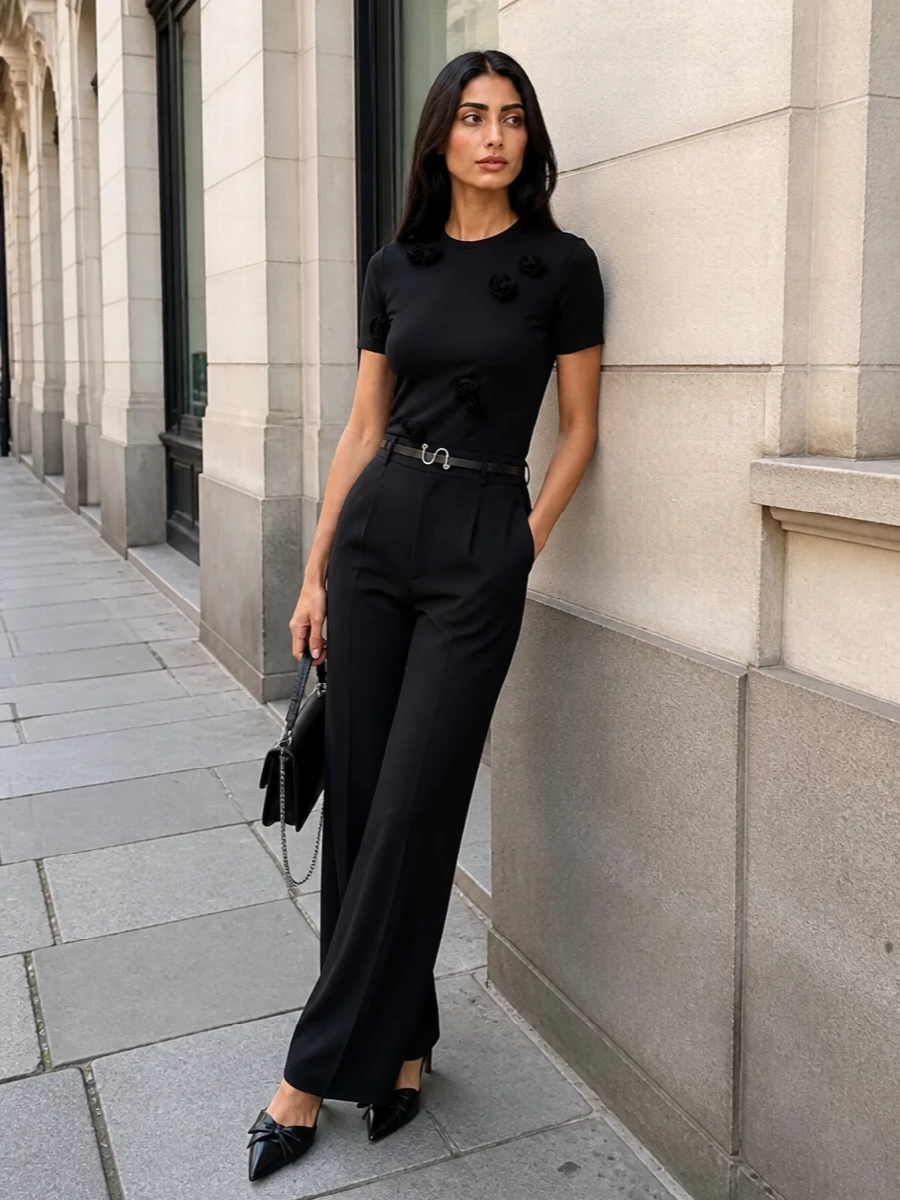 A sculpted black floral tee is grounded by high-rise, fluid wide-leg trousers for a long, modern column of monochrome tailoring. Pointed black slingback stilettos add romantic sharpness, while a slim silver-chained shoulder bag and discreet Saint Laurent belt keep the look edited, evening-ready and quietly luxurious.