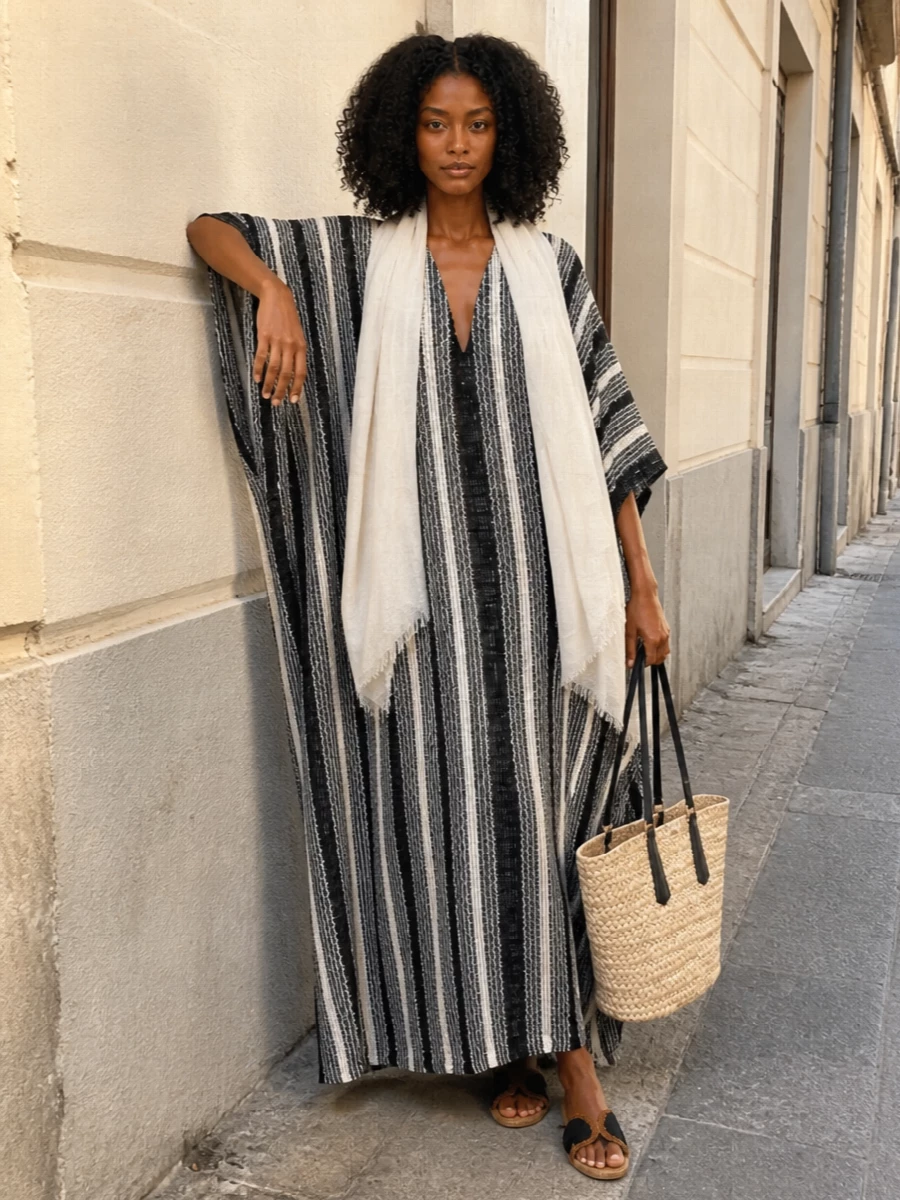 A monochrome striped kaftan anchors the outfit in graphic ease, offset by flat black espadrille slides that keep the mood grounded and relaxed. A structured straw tote with black leather handles ties into the palette while adding just enough polish, and a gauzy ivory wrap softens the lines for sun-faded terraces or slow city afternoons.