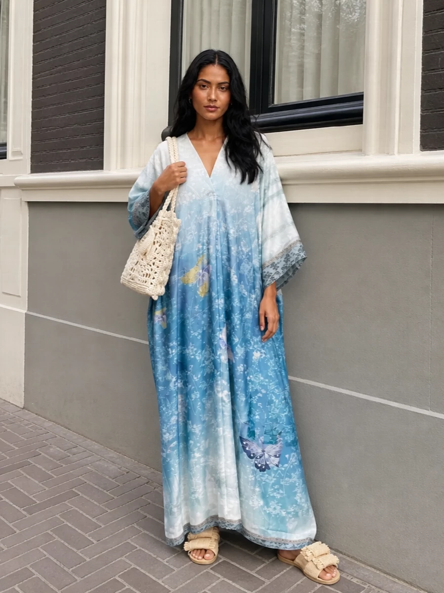A sky-washed Pierre Louise Mascia kaftan takes center stage, its butterflies and ombré blues drifting around the body like painted light. Natural raffia slides and an ivory crochet tote keep the mood barefoot and sunlit, matching the dress’s softness without adding weight. The result reads like a summer daydream, ideal for long lunches by the water or artful resort evenings on warm stone terraces.