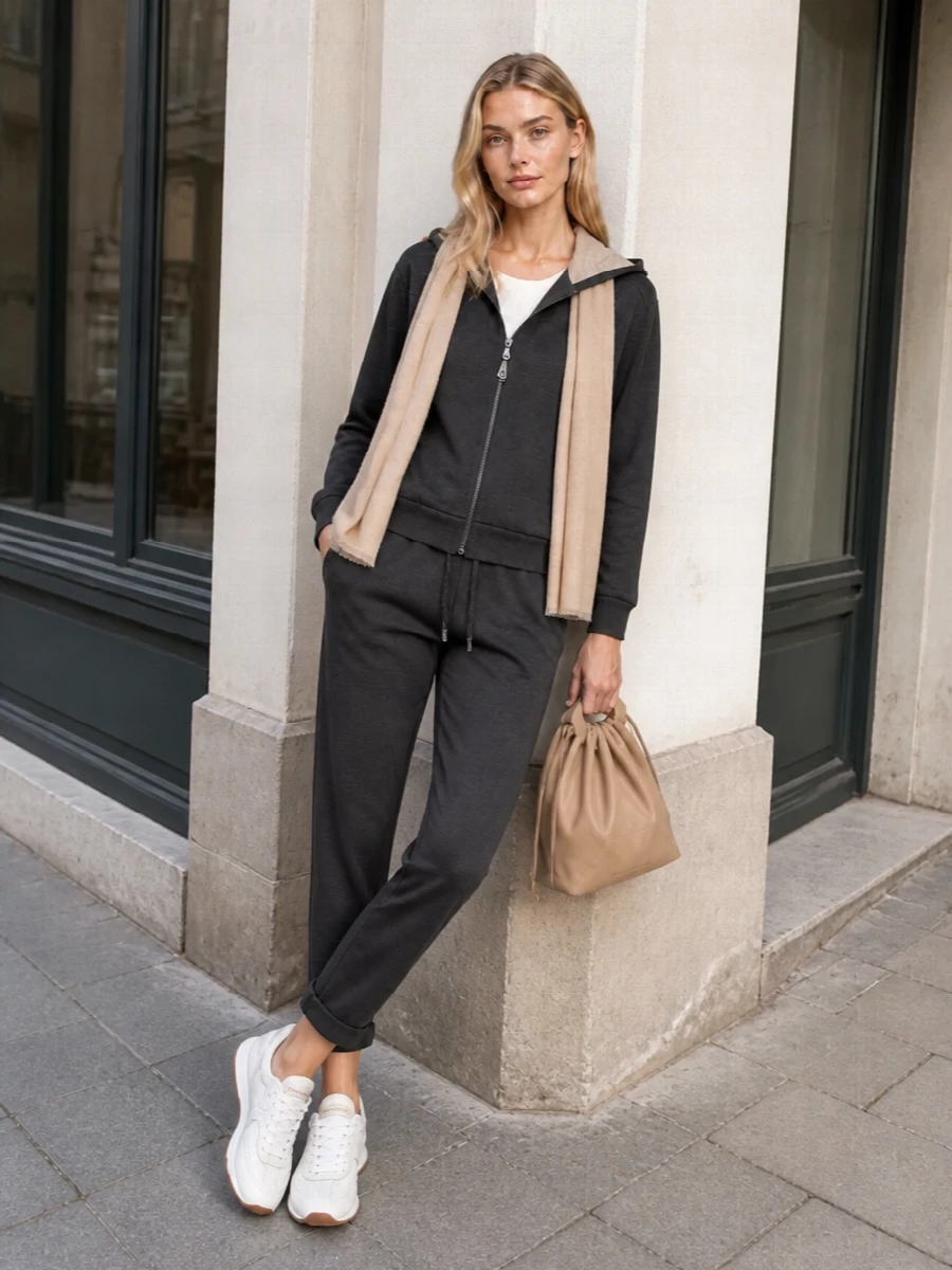 This look frames the charcoal Brunello Cucinelli hoodie with its matching tapered jogger for a refined, monochrome sport-luxe set that feels polished enough for city days yet effortless for travel. Soft white runners brighten the base while a taupe-beige drawstring pouch and airy sand scarf introduce warm, tonal accents that echo the hoodie’s lining and keep the palette cohesive and quietly luxurious.