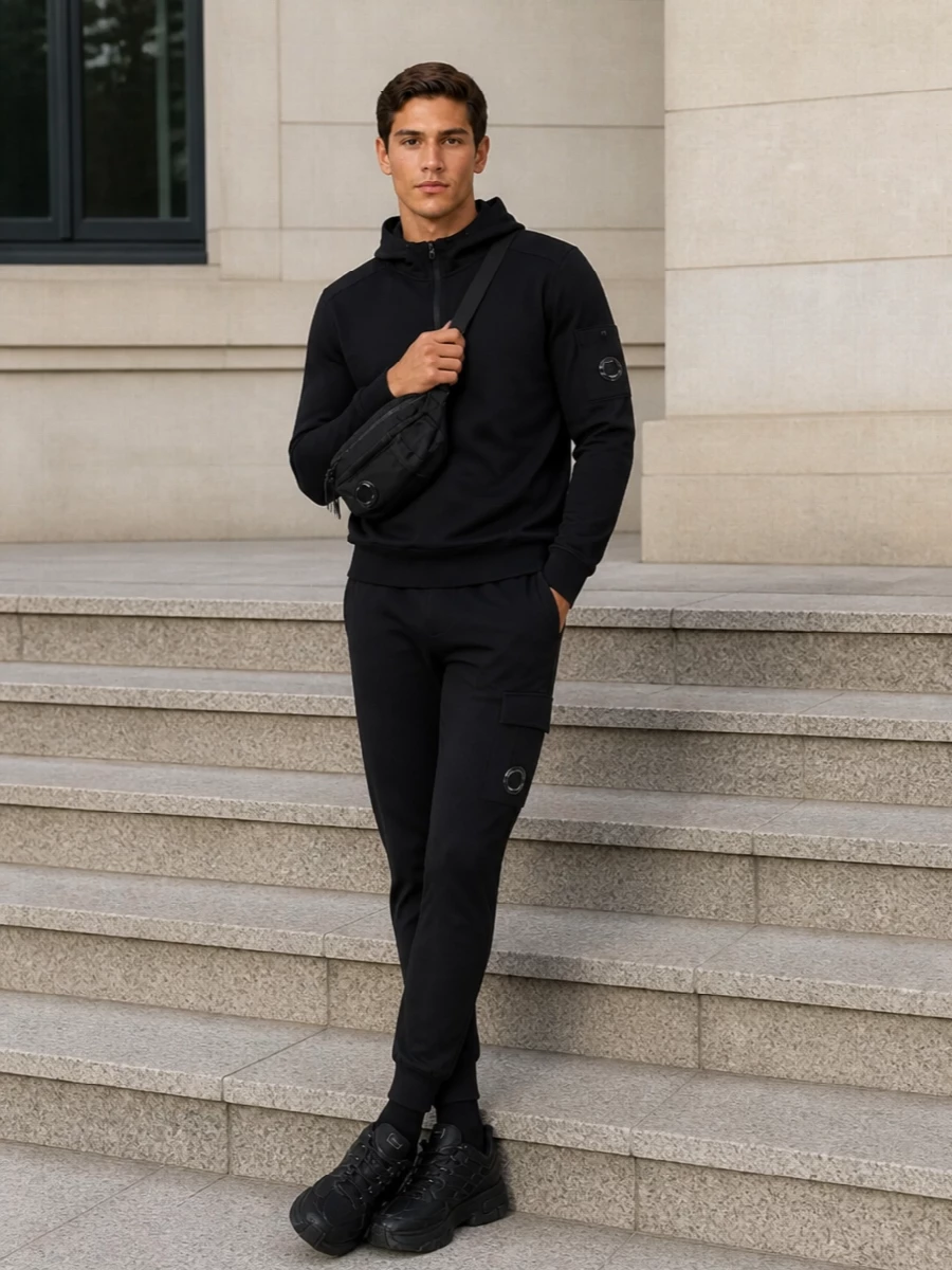 A stealthy C.P. Company uniform built around black technical joggers and a matching half‑zip hoodie, unified by lens details and clean lines. Chunky all‑black Salomons and a compact crossbody keep it ready for city miles, while ribbed black socks finish the silhouette with a quiet, minimal edge. The result is a focused, monochrome streetwear set that feels engineered for movement and travel.