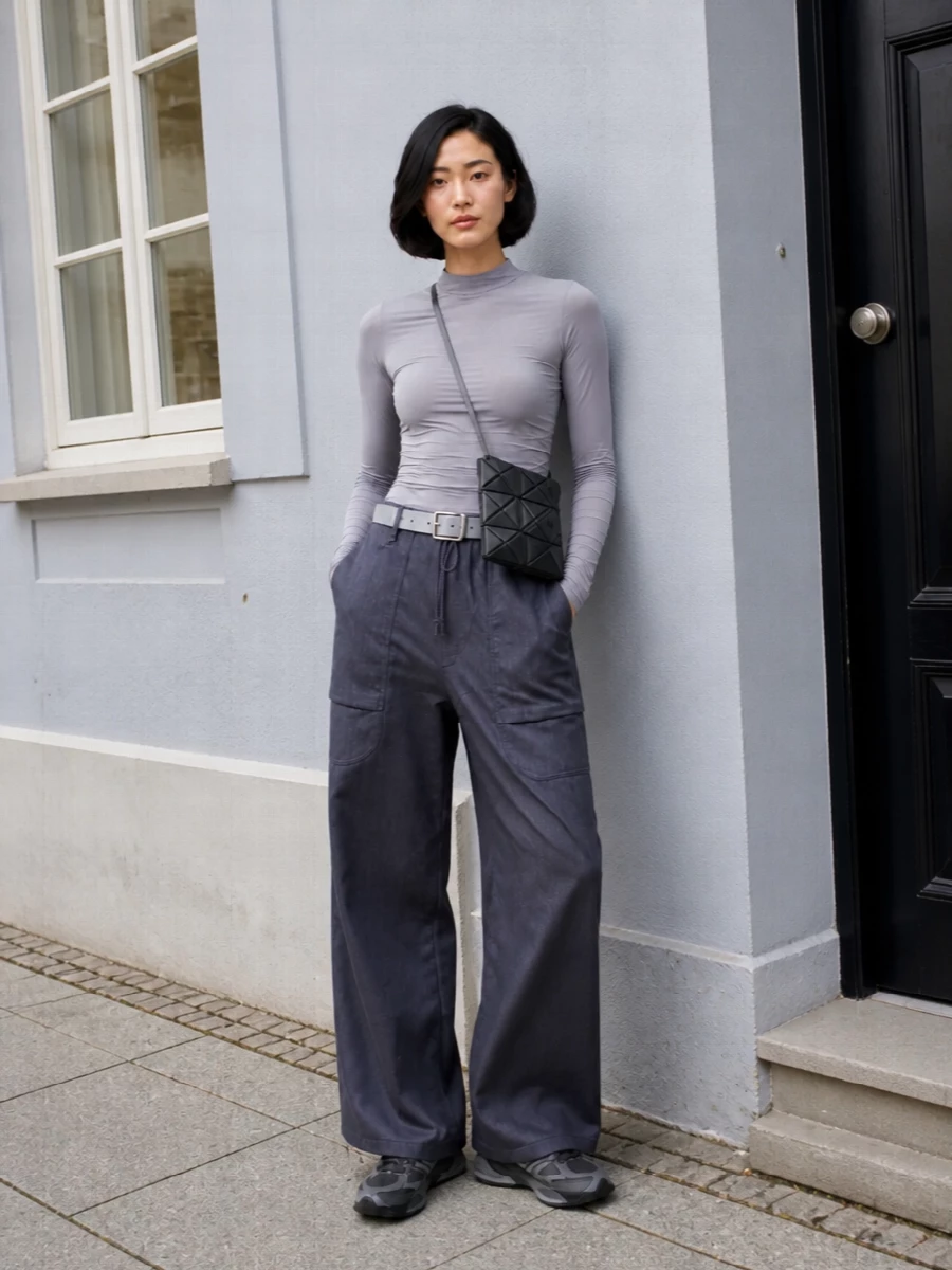 Ink navy volume on the bottom is balanced by a slim, ruched grey top and a run of tonal greys through sneakers and accessories. The result feels like a quiet, technical uniform, all about proportion and texture rather than logos. Black geometric hardware in the bag keeps the mood urban and precise without interrupting the soft, cool palette.