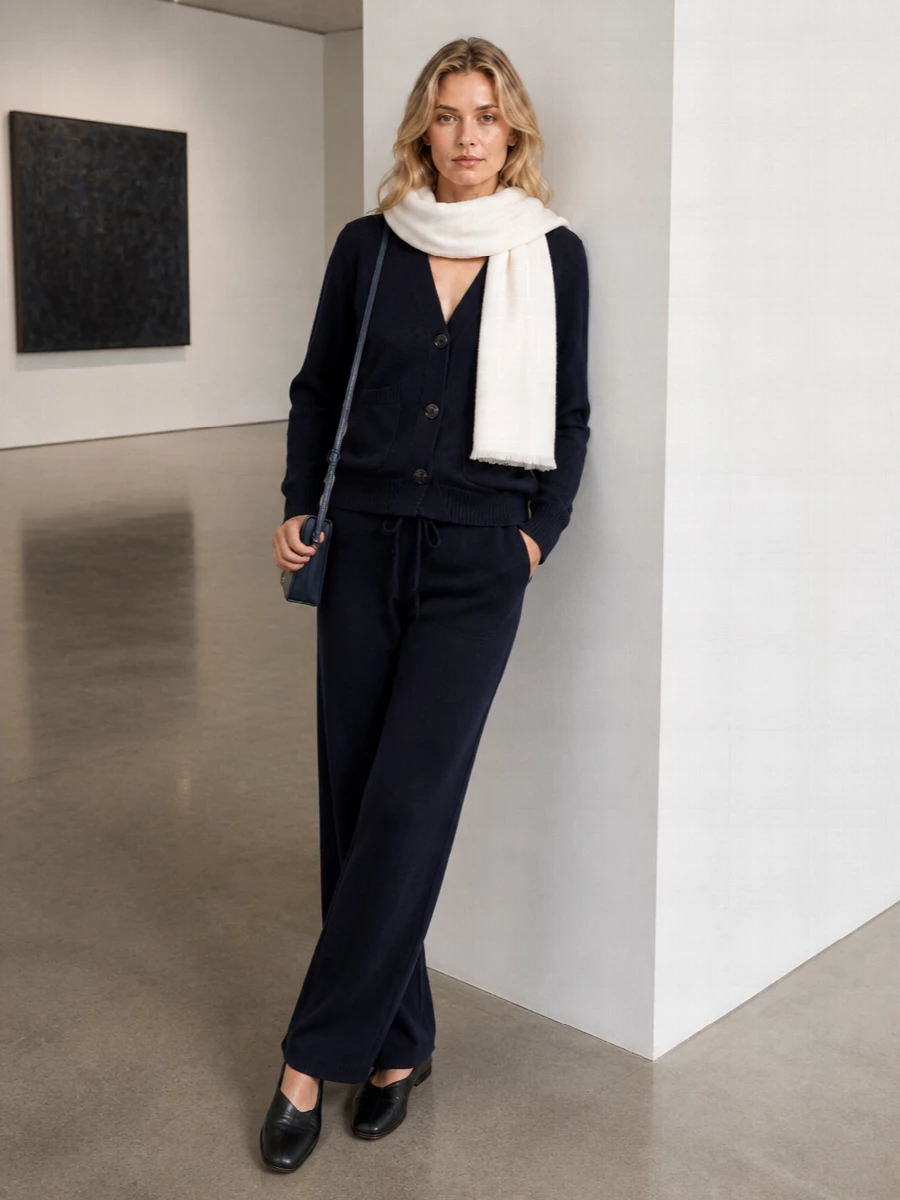 A navy-on-navy knit column keeps the mood calm and precise, with the wide-leg pant elongating the line under the relaxed V-neck cardigan. Black sculptural flats and a slim navy crossbody sharpen the softness, while an oversized ivory scarf adds a quiet halo of light around the face. The result is a quietly luxurious winter uniform that moves easily from home to low-key city days without losing its restraint.