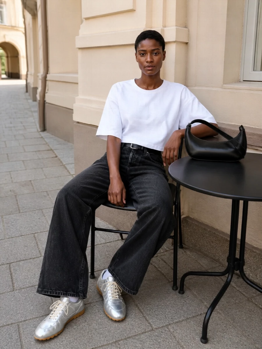 High-rise washed-black Haikure denim sets a strong, wide-leg base framed by a cropped, boxy white tee that lands right on the waistband. A slim black belt crisps up the waist, while metallic silver sneakers and a curved black hobo bag keep the palette tight and modern. The result is a clean, city-ready uniform that feels minimal, directional and easy to wear all day.