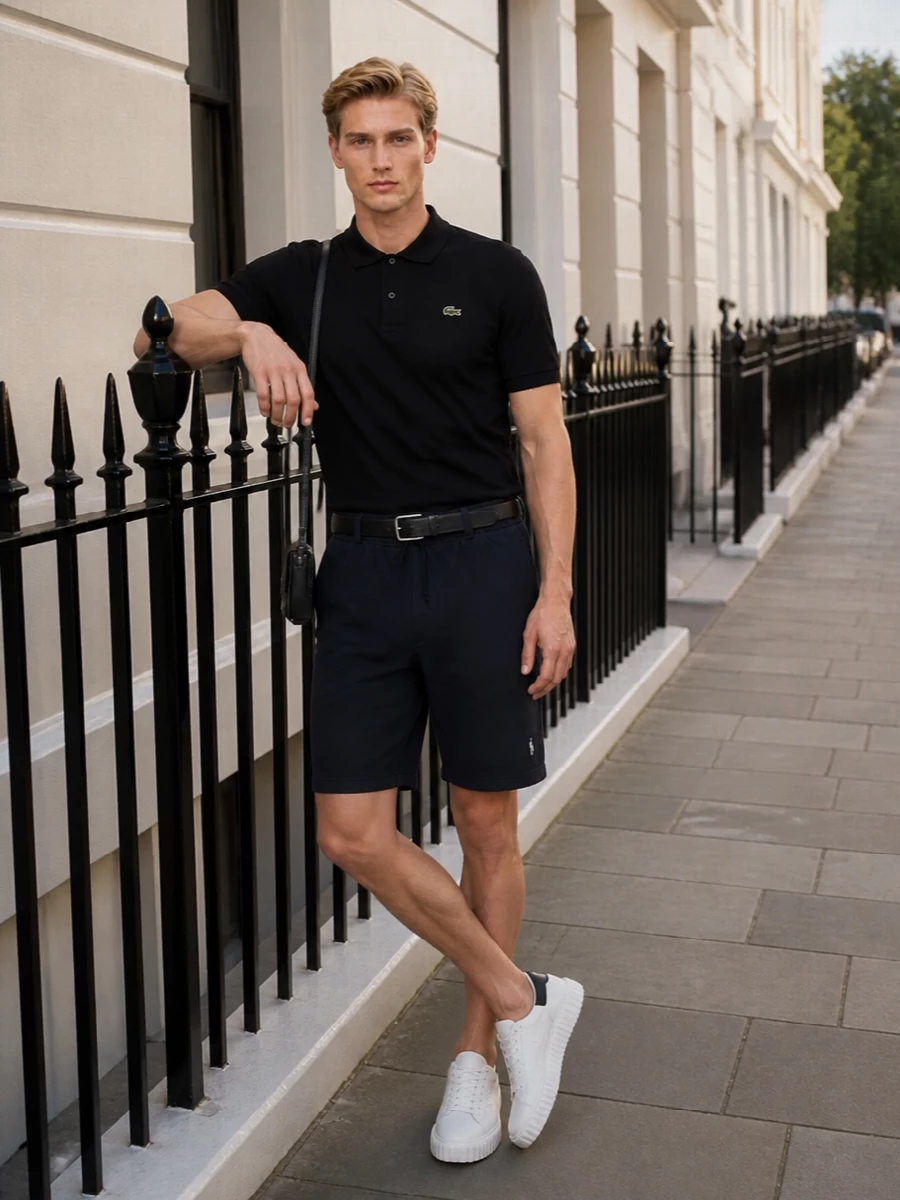 A black Lacoste polo sits over deep navy drawstring shorts for a clean sport-prep uniform that feels relaxed but still intentional. White leather sneakers and a slim black shoulder bag keep the lines light and sharp, while a discreet leather belt adds just enough structure at the waist. It is an easy warm-weather combination that works for city days, terraces and travel alike.
