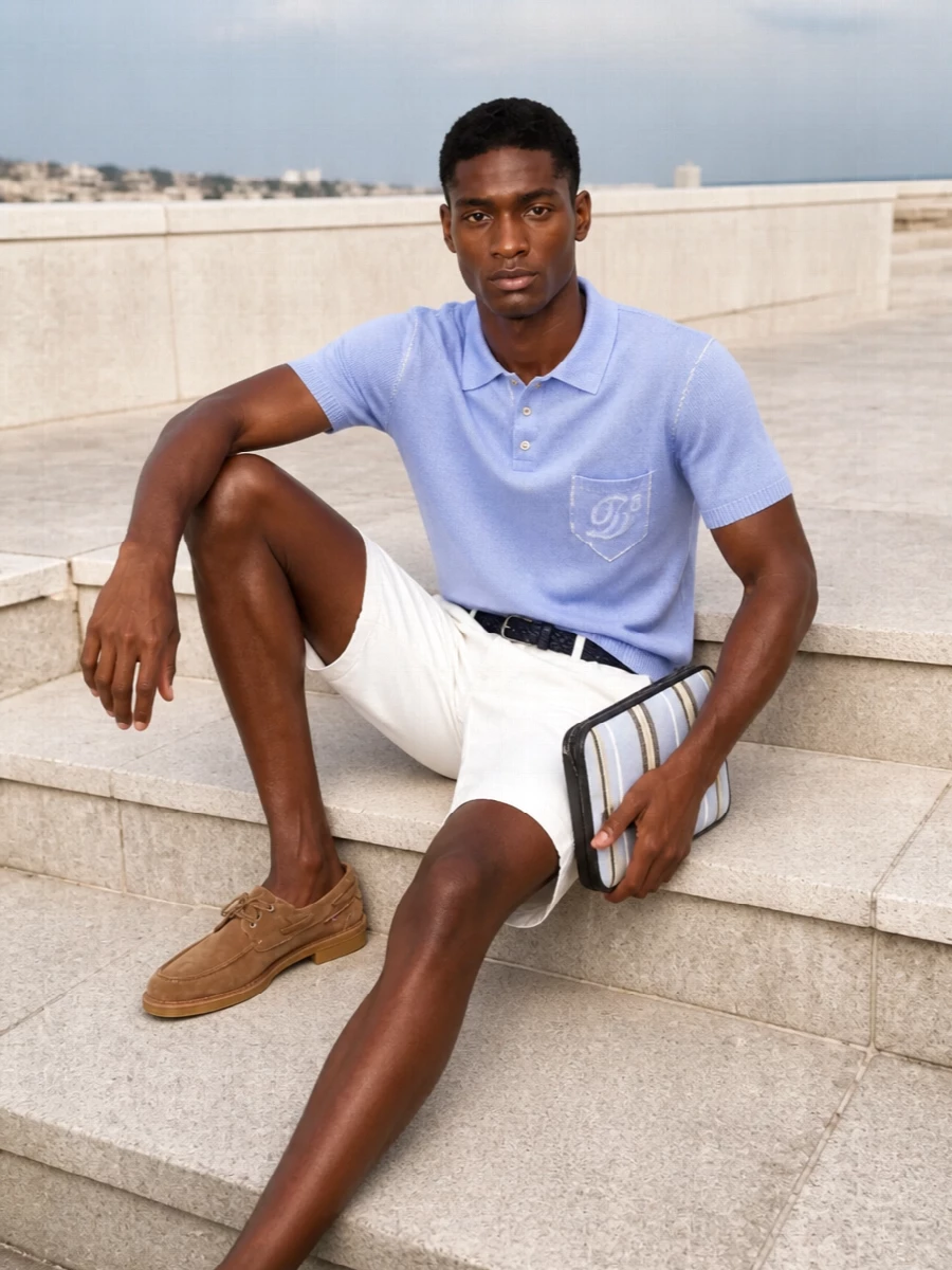 Soft white Bermudas meet an ice blue knit polo for a clean, preppy take on warm weather dressing. Tan suede moccasin loafers and a striped canvas clutch pull in sunlit coastal tones, while a navy braided belt gives just enough structure at the waist. The result is relaxed but precise, ideal for city terraces or a long lunch by the water.