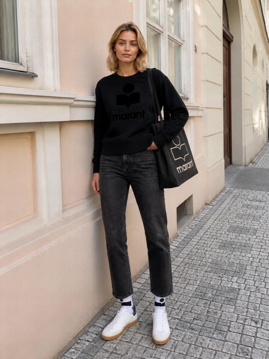 A black logo sweatshirt sits over washed charcoal denim for a tight, monochrome take on Marant’s off-duty uniform. White leather sneakers and a structured logo tote sharpen the streetwear mood without losing ease. Graphic socks peeking between hem and sneaker keep the whole mix feeling considered but still low-key.