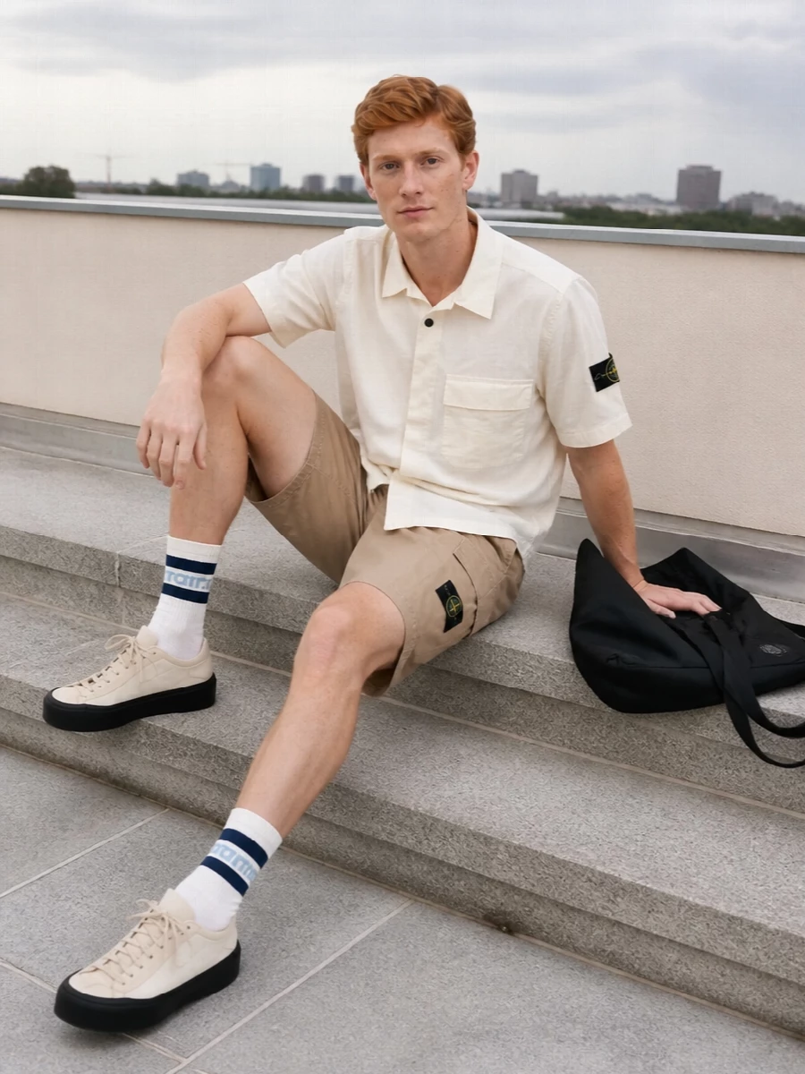 A light Stone Island cargo short paired with an off-white utility shirt keeps the palette crisp and neutral for warm urban days. Technical low-top sneakers and an oversized black tote ground the sand tones with a subtle street edge. Soft striped socks peek out just enough to hint at sport without distracting from the clean, uniform feel.