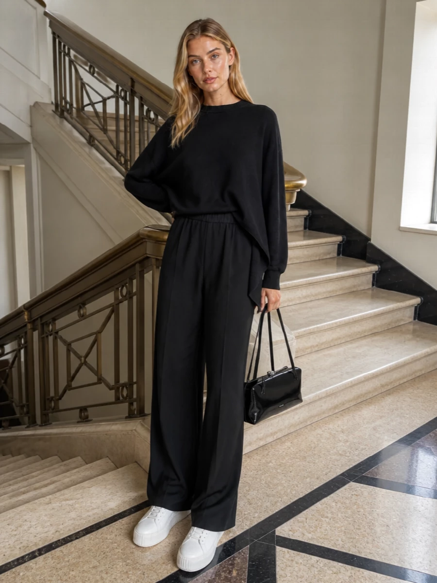 An all-black column anchors the asymmetric sweatshirt in a way that feels calm, sculptural and quietly confident. Fluid wide-leg trousers and a glossy structured shoulder bag keep the mood polished yet relaxed, while bright white platform sneakers add a precise, modern offset without disrupting the minimalism.