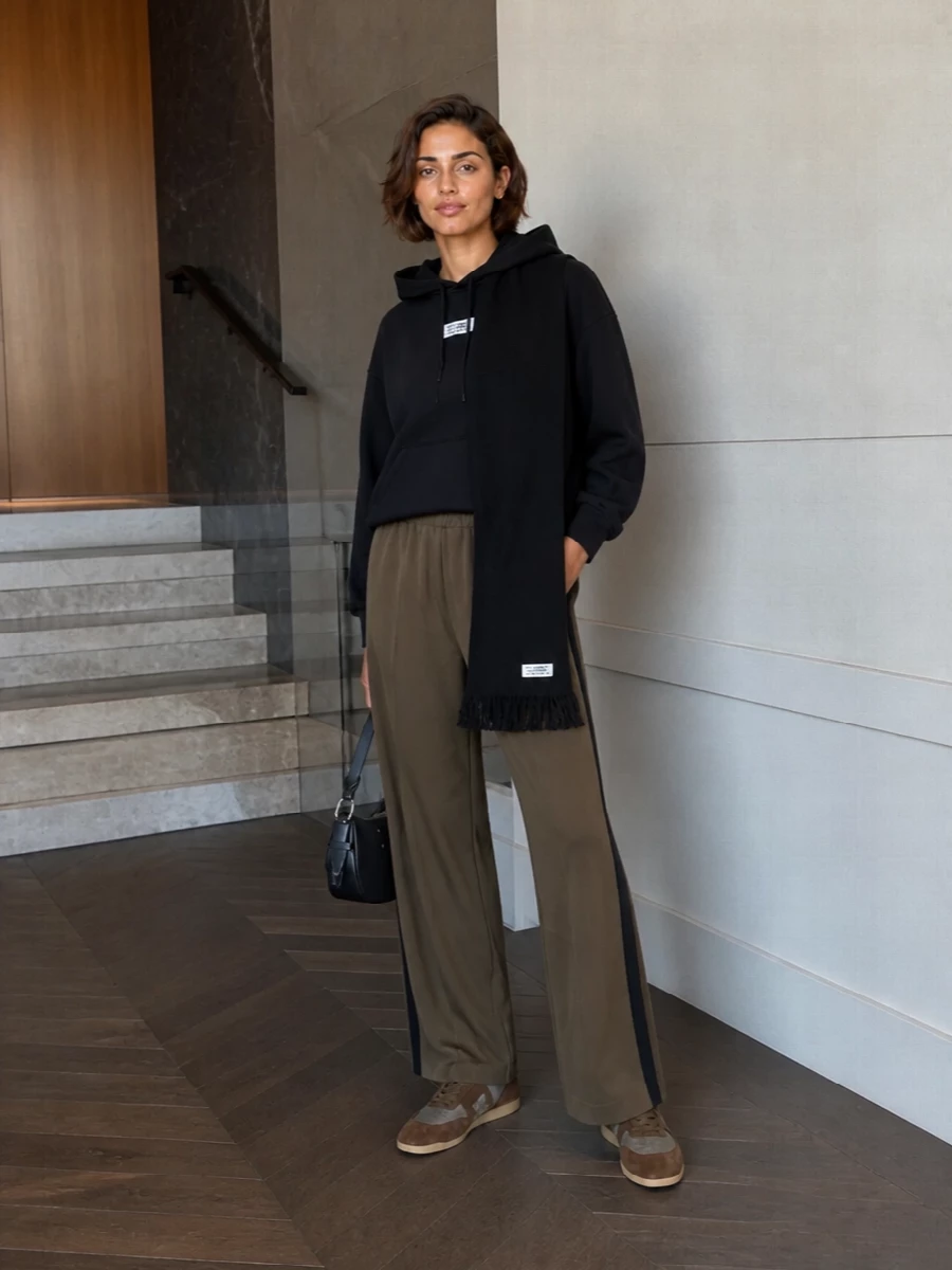 Olive track pants meet a cropped black hoodie for a relaxed, Margiela-coded take on city sport. Tonal brown sneakers keep the palette grounded, while a curved black shoulder bag and ribbed scarf sharpen the finish without breaking the ease.
