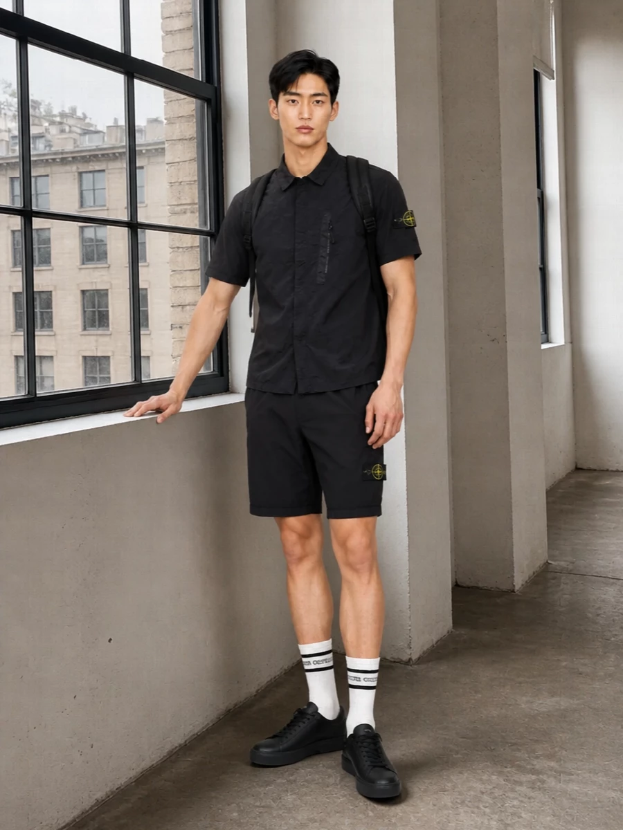 A head-to-toe Stone Island uniform in black technical fabrics keeps the energy clean, functional and street-ready. The boxy nylon shirt sits just off the regular-fit cargo shorts, so the set feels intentional rather than matchy, with the all-black sneakers and backpack tightening everything into a sharp, city-focused kit. Graphic striped socks add the only soft contrast, giving a discreet nod to sport without breaking the monochrome line.
