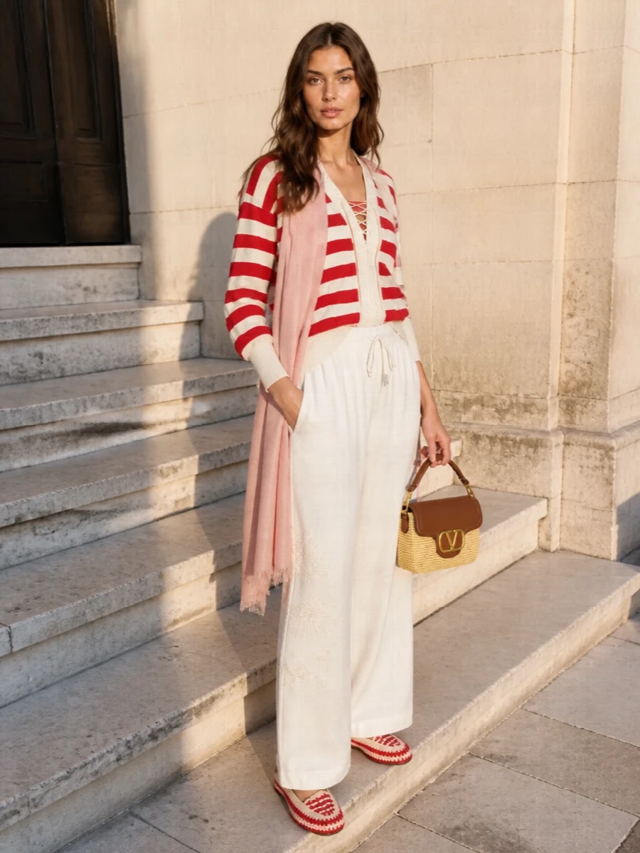 A relaxed red-and-cream striped cardigan is grounded with airy embroidered wide-leg trousers for a soft, resort-ready silhouette that still reads polished. Red crochet flats pick up the maritime stripes, while a tan raffia shoulder bag and blush stole bring in sun-washed neutrals that keep the palette warm and refined. The mix feels quietly coastal and put-together, perfect for days that drift from seaside lunch to terrace drinks.