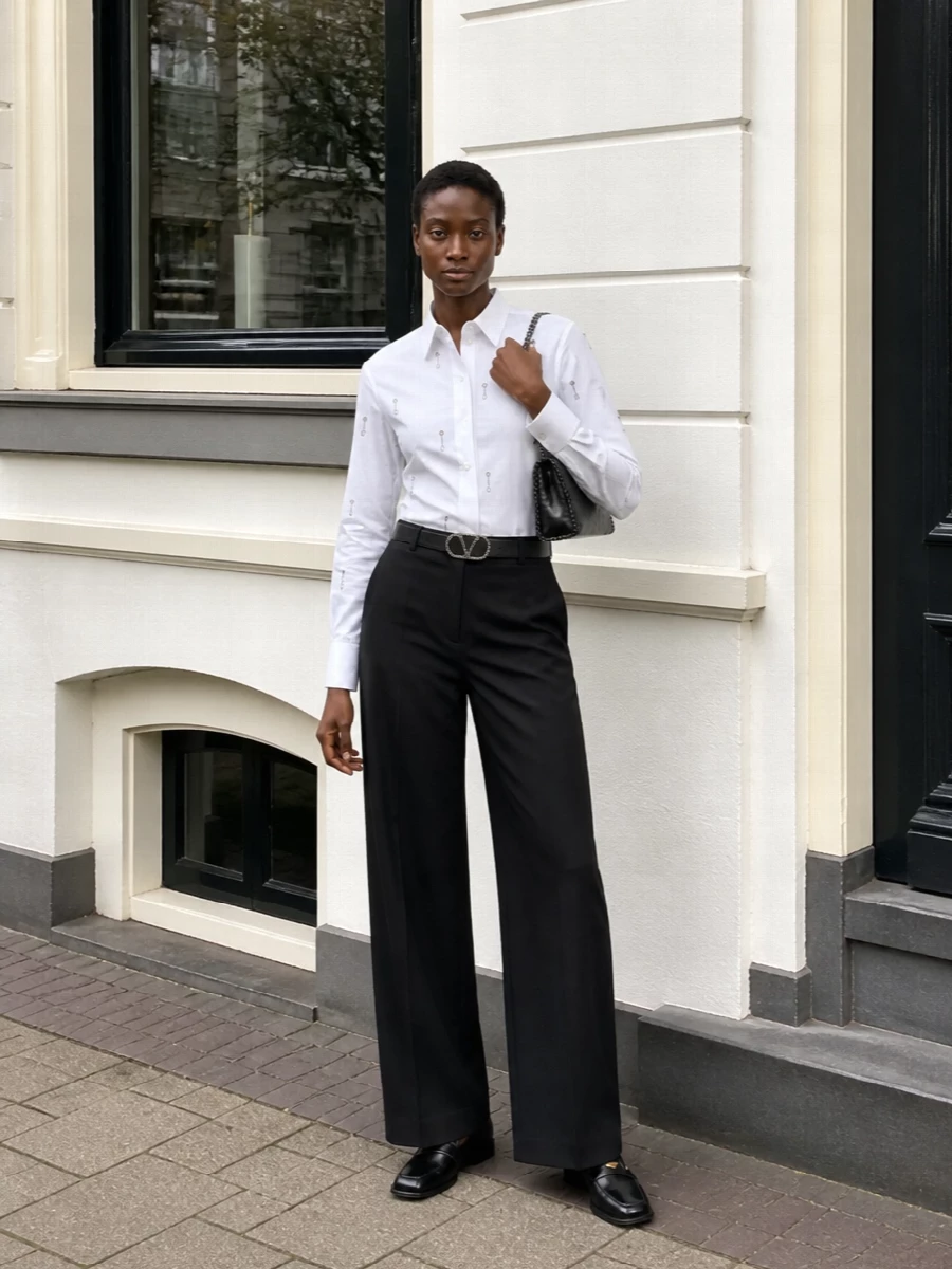 Stella McCartney’s crystal-pinned shirt is grounded with impeccably cut high-rise black wide-leg trousers for a long, modern line. A structured loafer keeps the mood sharp rather than overtly evening, while a chain-trim clutch and crystal-detailed belt quietly repeat the metallic accents for a cohesive, boardroom-to-dinner finish.