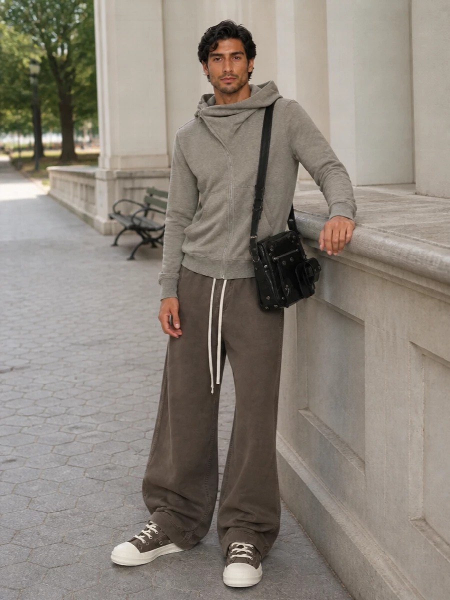 This look leans into a soft, dystopian monochrome: the washed taupe hoodie sits over gravity-heavy ultra-wide trousers in a closely related taupe-brown, creating one long, fluid column of Rick volume. Taupe high-top sneakers continue the neutral story and balance the pooling hem, while a compact black crossbody injects just enough hardware and contrast to keep the outfit sharp and urban without disrupting its quiet, utilitarian ease.