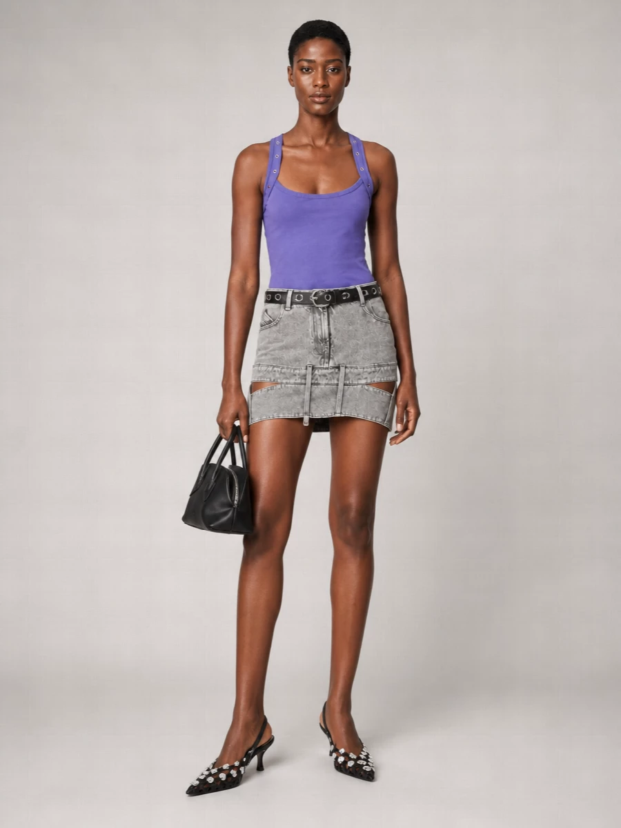 A saturated purple tank tops a cut-out grey denim mini for a tight, attitude-heavy Y2K silhouette. Black studded kitten heels and a structured mini bag keep the palette focused and hardware-driven, while a slim eyelet belt crisps the waist and amplifies the tank’s grommeted straps. The result is a compact, night-ready city uniform that feels bold without losing ease.