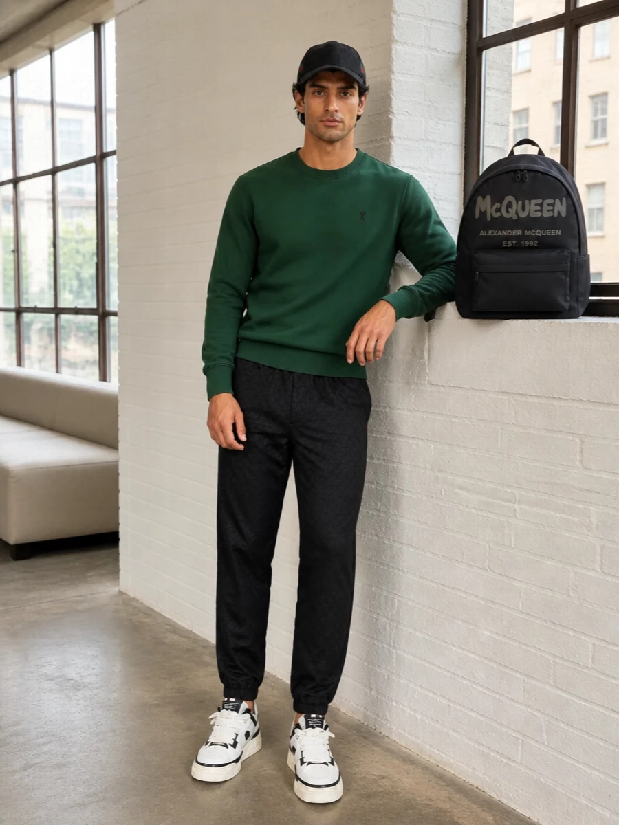 A deep forest-green sweatshirt anchors the outfit against sharp black jacquard track pants for a clean, athletic base that syncs with the Gucci cap’s quiet branding. Chunky off-white and black Amiri sneakers add volume and sport energy at the ground, while the logoed McQueen backpack keeps the whole combination firmly in luxe streetwear territory. It feels ready for city days that move from errands to late-night hangs without ever looking overdone.