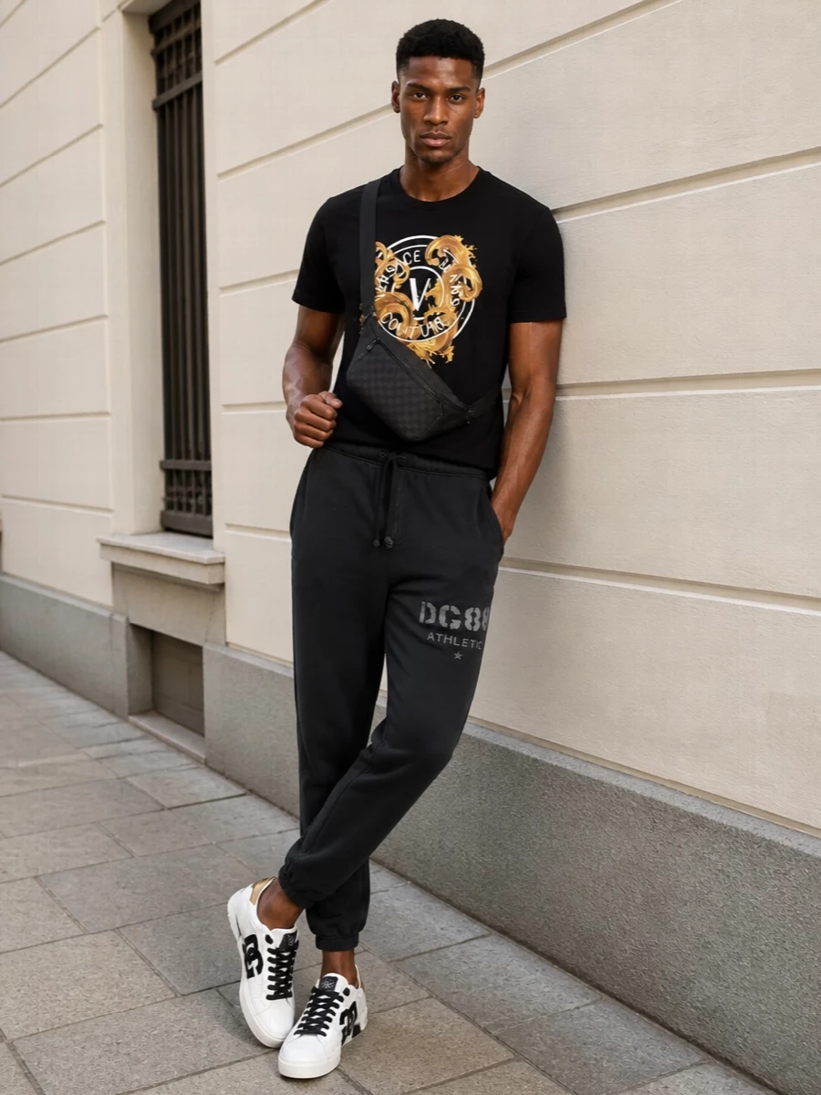 A bold Versace tee anchors this outfit in black and gold, paired with charcoal joggers that keep the silhouette relaxed and sport-led. White and gold-accented sneakers brighten the base while echoing the graphic branding, and a sleek black Gucci sling bag adds just enough structure without breaking the streetwear mood.