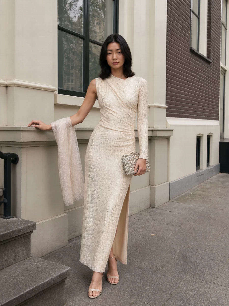 A softly luminous ivory column is paired with barely-there champagne sandals to keep the line long and fluid. A crystal-splashed minaudière and gossamer wrap add just enough shimmer, giving cocktail or wedding evenings a polished, romantic finish without crowding the asymmetric neckline.