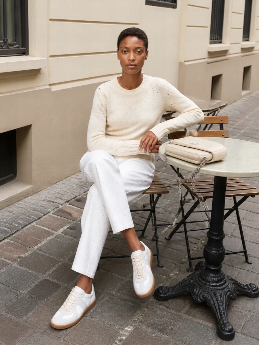 A buttercream-on-ivory palette keeps the distressed knit and paint-speckled trouser feeling intentional rather than undone, reading like a uniform for someone who lives between studio and street. Off‑white sneakers and a padded buttercream bag stay within the same whisper-soft spectrum, so the numeric-print scarf becomes a quiet focal point instead of competing with color. The result is relaxed, minimal and quietly clever, grounded in Margiela’s art-school precision.