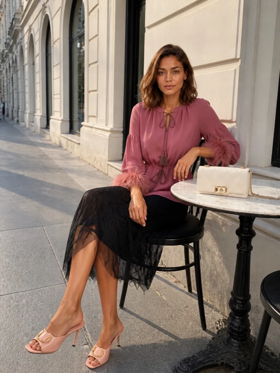 A dusty-rose tasselled blouse meets dark romantic lace for a cocktail outfit that feels soft, modern and assured. The black asymmetric skirt sharpens the silhouette, while blush heels and an ivory quilted bag keep the palette luminous around the face and legs. Gold accents quietly echo throughout, so the feathers and texture do the talking without the styling ever feeling busy.