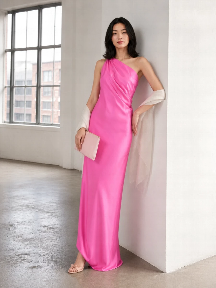 A vivid satin column in liquid pink holds all the drama, so everything around it stays pared back and precise. Barely-there blush heels and a slim pink clutch keep the palette whisper-soft, letting the asymmetric neckline and fluid length read as the statement. An airy ivory stole finishes the composition with a light, romantic veil that still feels resolutely modern.